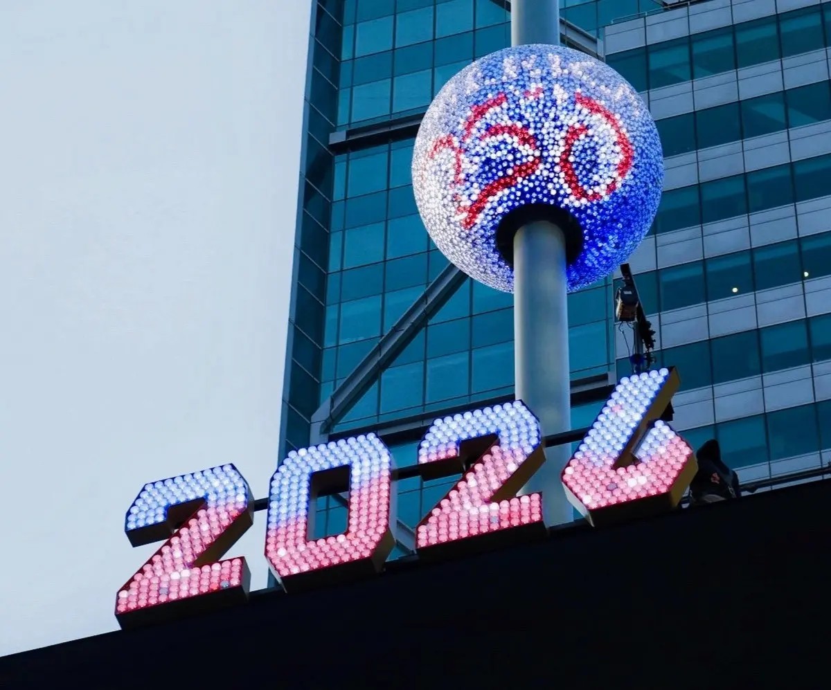 This is a Times Square Ball drop for America 250. A large illuminated sign showing the numbers 2024 glows in red, white, and blue lights atop a building. Above the numbers is a spherical structure covered in small lights, displaying the America 250 logo in red and blue. The sphere is mounted on a tall pole. A modern glass high‑rise building fills the background, with reflections visible in its windows. The sky appears overcast.