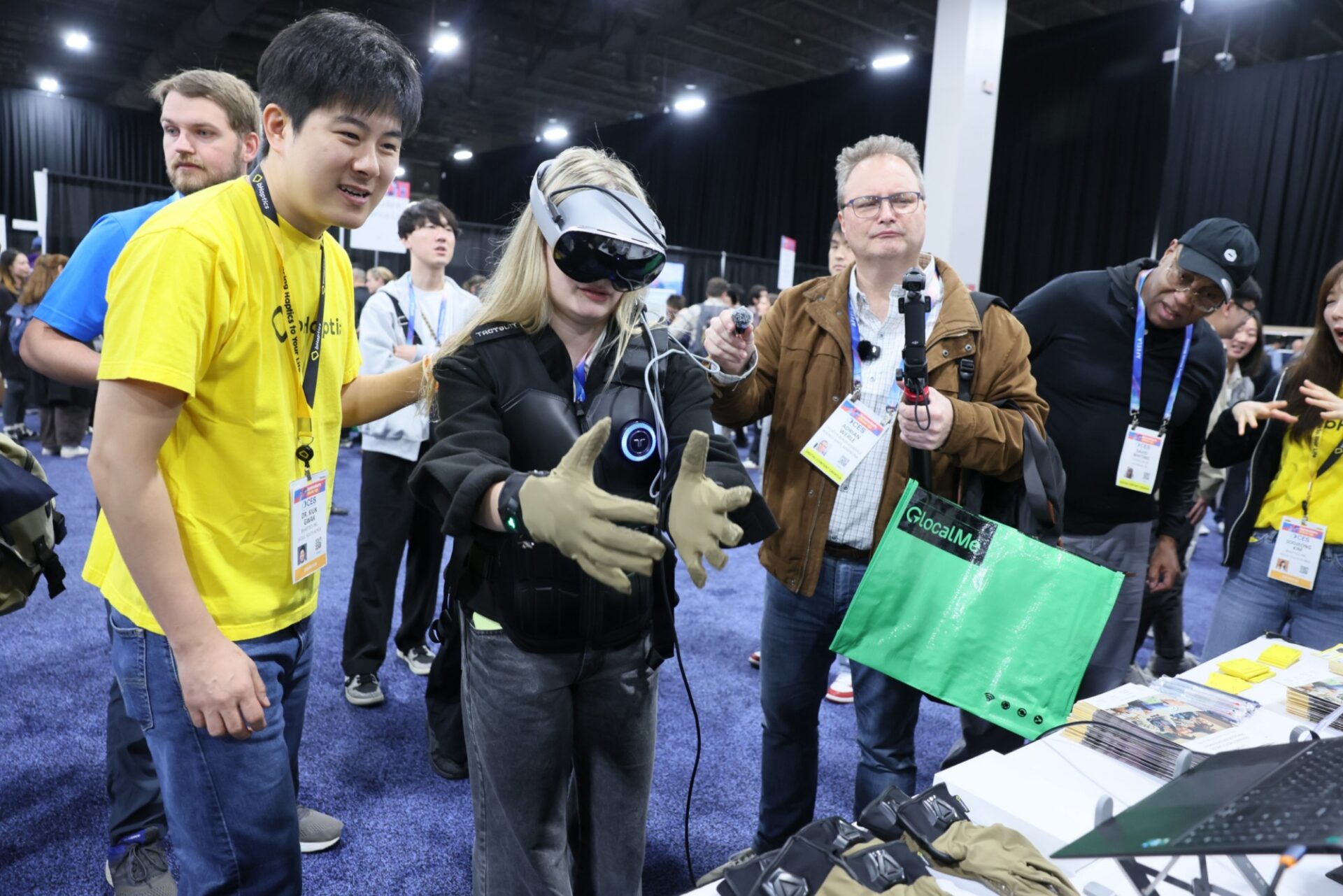 A person in the center of the image is wearing a virtual reality headset, padded gloves, and a vest with circular electronic components on the front. The individual is standing with arms extended while connected cables run from the equipment. Surrounding this person are others wearing event badges and casual clothing. One individual on the right is holding a handheld device with a mounted camera or sensor. Another person on the left is wearing a bright yellow shirt. The background shows a large indoor event space with carpeting, overhead lighting, and other attendees moving around. A green tote bag labeled GlocalMe is hanging from one person’s shoulder. A table in the foreground displays additional padded gear or equipment.
