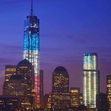 A nighttime city skyline featuring One World Trade Center and tall modern buildings illuminated against a deep blue and purple sky. The tallest skyscraper in the center is lit from bottom to top in red, white, and blue horizontal sections. Additional buildings on either side also display vertical bands of bright white and blue lights. The scene highlights an urban landscape with reflective windows and distinctive rooftop shapes.