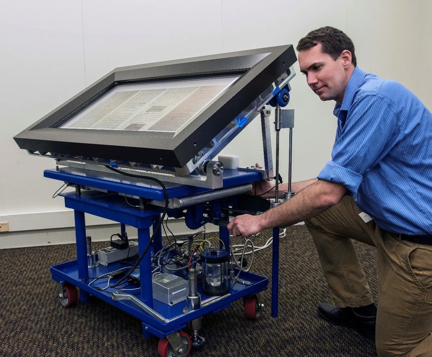 A person is kneeling beside a large mechanical device that appears to be a scanning or imaging machine. The device has a rectangular glass surface held within a metal frame, positioned at an angle, with a page or document visible underneath the glass. The machine is mounted on a blue wheeled platform with multiple exposed components, including wiring, metal supports, hydraulic or pneumatic cylinders, and various electronic parts. The individual is adjusting or examining a component beneath the angled surface. The scene takes place in an indoor workspace with a neutral-colored wall and carpeted floor.