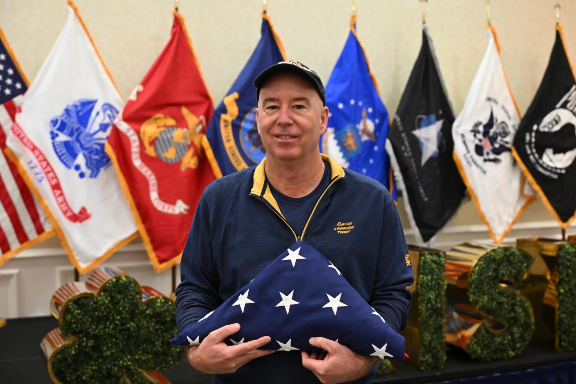 A person stands indoors holding a folded American flag in both hands. Behind the individual is a row of military service flags displayed on stands, including flags for different branches of the United States Armed Forces. The person is wearing a dark long‑sleeved top with a zippered collar and a cap. In the foreground, decorative elements shaped like letters are partially visible, covered in green foliage. The setting appears to be a ceremonial or commemorative event.