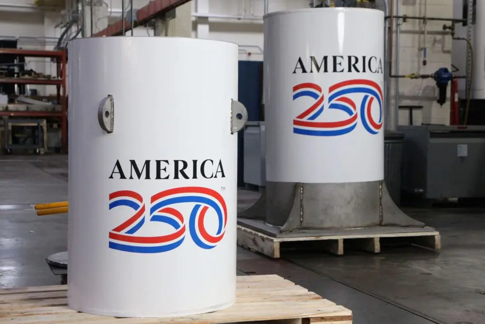 Nist A250 Time Capsule 003 Slide Cr The image shows two large cylindrical metal containers positioned indoors on a warehouse or workshop floor. Both containers are painted white and prominently display the text “AMERICA 250” in bold lettering, with a stylized red and blue graphic forming the number “250.” The container in the foreground sits on a wooden pallet and has a smooth surface with small metal fixtures attached to its side. Behind it, a second, slightly taller container stands on a low platform with a metallic base. The surrounding environment includes industrial elements such as pipes, machinery, and shelving in the background, suggesting a manufacturing or storage setting. The lighting is even and bright, highlighting the clean surfaces of the containers and the crisp, patriotic colors of the printed design.